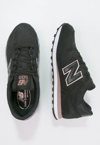 Svarta New Balance-sneakers har en överdel i mockasin, tygfoder, vit mellansula, gummiyttersula och en framträdande silverfärgad "N"-logotyp på sidorna.