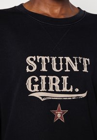 Svart sweatshirt med texten "STUNT GIRL" tryckt i sliten vit stil. Har en röd stjärngrafik med "PINKO" text längst ner.