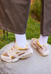 Beiga och bruna sneakers med nätpaneler, gula accenter och synlig luftdämpning. Vita sockar i kombination med ljusgrå byxor.