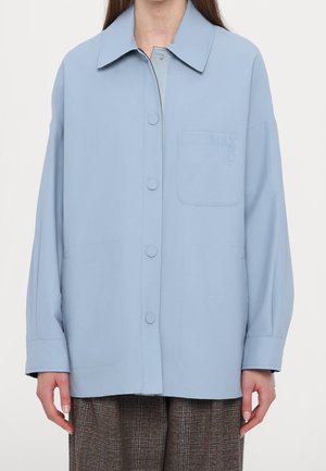 Vrouw draagt een losse lichtblauwe blouse met knopen, kraag en borstzak, gecombineerd met bruine geruite broek tegen een witte achtergrond.
