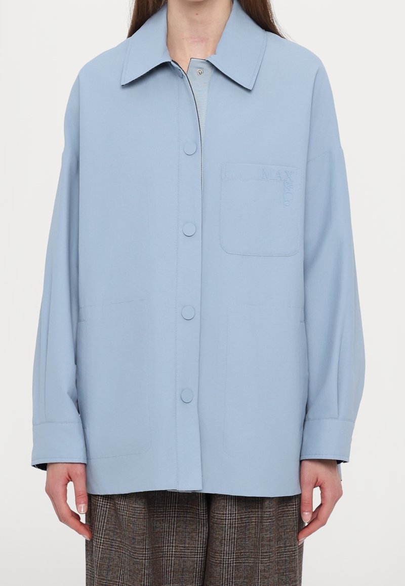 Femme portant une chemise ample bleu clair à boutons avec col et poche poitrine, associée à un pantalon à carreaux marron sur fond blanc.