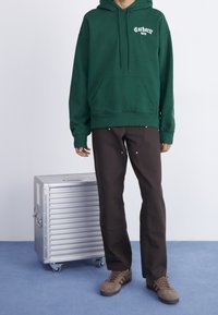 Sudadera con capucha verde oscuro con bolsillo frontal, de corte amplio. Pantalones marrones con detalles metálicos dorados. Zapatos grises. Maleta metálica con ruedas cerca. Fondo de piso azul.