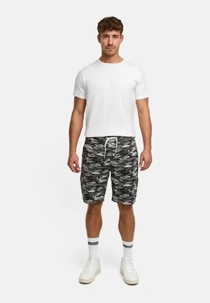 Mand, der står fremadrettet iført hvid t-shirt, sorte og grå camouflage-shorts, hvide sokker med sorte striber og hvide sneakers.