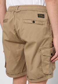 Khaki cargoshorts met een elastische tailleband, zijzakken en een label op de achterkant. Katoenen materiaal, opgerolde zoom en stevige stiksels.