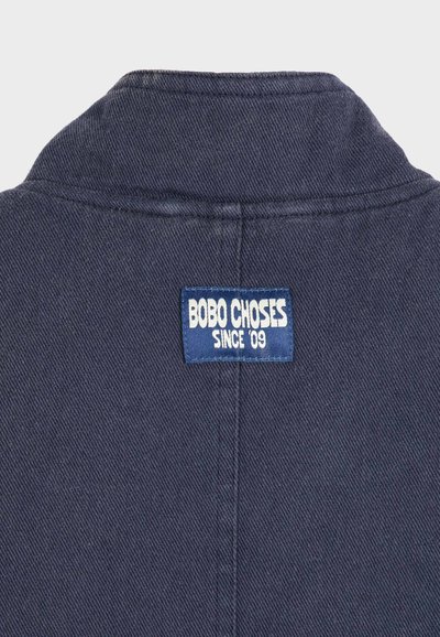 Tissu bleu foncé avec un col plié et une étiquette rectangulaire bleu marine portant l'inscription "BOBO CHOSES SINCE '09" cousue au centre du dos.