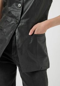 Svart läderväst med skräddarsydd passform, utrustad med två knappar, en sidoficka och slät yta. Kombinerad med svarta denimbyxor.