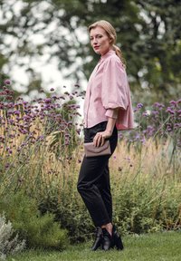 Light pink puff-sleeve blouse, black tapered pants, and black ankle boots. Holding a taupe wallet with a gold accent. Surrounded by flowers.
