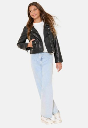 Nuori tyttö, joka pukeutunut mustaan nahkatakkiin, valkoiseen paitaan, vaaleansinisiin farkkuihin ja valkoisiin tennareihin, seisoo käsi takin taskussa.