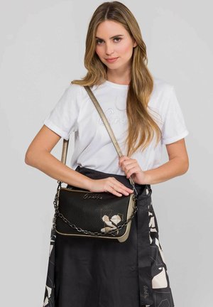 Mujer de cabello largo con camiseta blanca y falda negra, sosteniendo un bolso de hombro negro y beige con detalle de cadena y diseño abstracto.