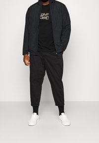 Černá lehká bunda přes černé tričko s logem "Calvin Klein", doplněná černými zúženými kalhotami a bílými teniskami. Jednoduchý, moderní design.