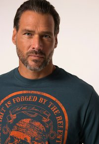 Camiseta de manga larga verdeazulada con un llamativo estampado gráfico naranja que incluye texto y un diseño de calavera, hecha de suave material de algodón.