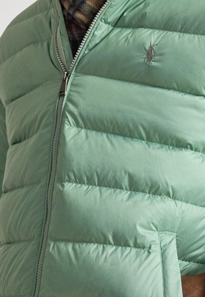 Zelená nadýchaná bunda s prešívaným vzorom, vybavená predným zipsom a logom na ľavej strane hrudníka. Mäkká, hladká tkanina s ľahkým pocitom.