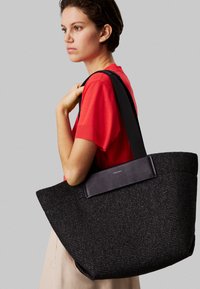 Borsa tote nera intrecciata con una superficie testurizzata, caratterizzata da un accentuato in pelle scura. La borsa ha manici larghi e una forma spaziosa e angolare.