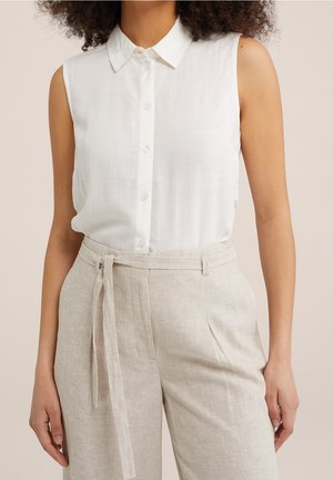 Femme portant un chemisier blanc sans manches à boutons, rentré dans un pantalon beige taille haute à ceinture nouée, debout, les bras détendus le long du corps.