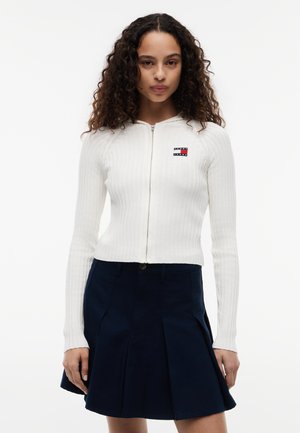 Žena s kučeravými vlasmi, oblečená v bielom rebrovanom svetri na zips s logom a námorníckou plisovanou sukňou na jednofarebnom pozadí.