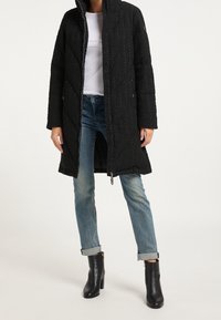 Sort quiltet lang frakke med høj krave, med lynlås foran og trykknapper; båret over en hvid top og blå jeans.