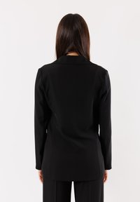 Blazer su misura nero con maniche lunghe, revers a punta, texture liscia e silhouette aderente. La vista posteriore enfatizza il design elegante.