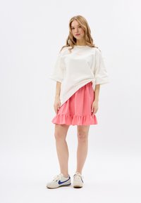Witte oversized t-shirt met roze letters, gecombineerd met een gerimpelde roze rok en beige sneakers met blauwe accenten. Gladde texturen.