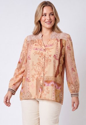 Vrouw die lacht en een perzikkleurige blouse met bloemenpatroon en kanten schouders draagt, met beige broek tegen een effen lichte achtergrond.