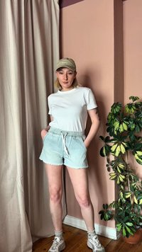 Νέα γυναίκα με λευκό μπλουζάκι, ανοιχτόχρωμα τζιν σορτσάκια, μπεζ καπέλο και αθλητικά παπούτσια στέκεται με τα χέρια στις τσέπες κοντά σε μια γλαστρική φυτό και κουρτίνα σε ένα ροζ δωμάτιο.