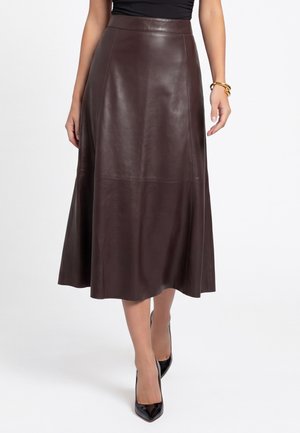 Vrouw draagt een donkerbruine leren midi-rok, zwarte puntige hakken, een zwart topje en gouden armbanden aan één pols.