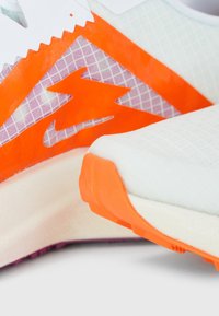 Gros plan sur une basket de sport blanche et orange avec un motif éclair orange translucide sur le côté et une semelle texturée.