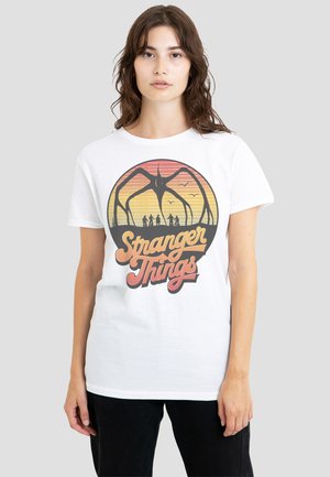 Young woman wearing a white T-shirt with a retro "Stranger Things" design featuring silhouetted kids and a large spider-like creature.