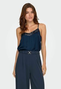 Marineblå satin camisole med tynde stropper, der har en halsudskæring pyntet med pailletter, parret med højtsiddende bukser og et dekorativt spænde bælte.