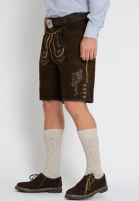 Ruskea mokkashortsit, joissa on monimutkainen brodeeraus, keltaisia korostuksia, sivupainikkeita, yhdistettynä vaaleisiin neulottuihin sukkahousuihin ja ruskeisiin nauhakenkiin.