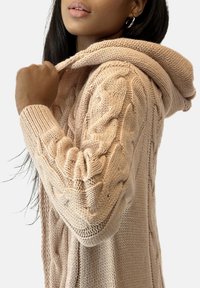 Ljusrosa stickad kofta med texturerat kabelmönster, huva och ribbade muddar, visad från sidan för att framhäva hantverk och detaljer.