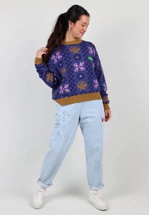 Sweatshirt crème à col rond de couleur violet avec des motifs floraux et solaires, bord côte marron. Jean baggy bleu clair avec des motifs gris complexes. Baskets blanches.