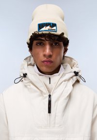Beanie beige con patch blu "NAPAPIJI" sul fronte e giacca bianca con cappuccio, dotata di tasca frontale, chiusura lampo nera e coulisse.