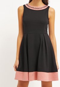 Svart ärmlös klänning med en åtsittande bodice och utsvängd kjol, med en rosa krage och rosa fåll. Dekorerad halsringning med prydnadsbeads.