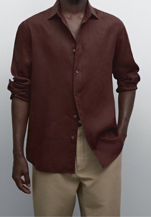 Homme portant une chemise marron foncé à manches longues déboutonnée avec les manches retroussées et un pantalon beige, debout avec une main dans la poche.