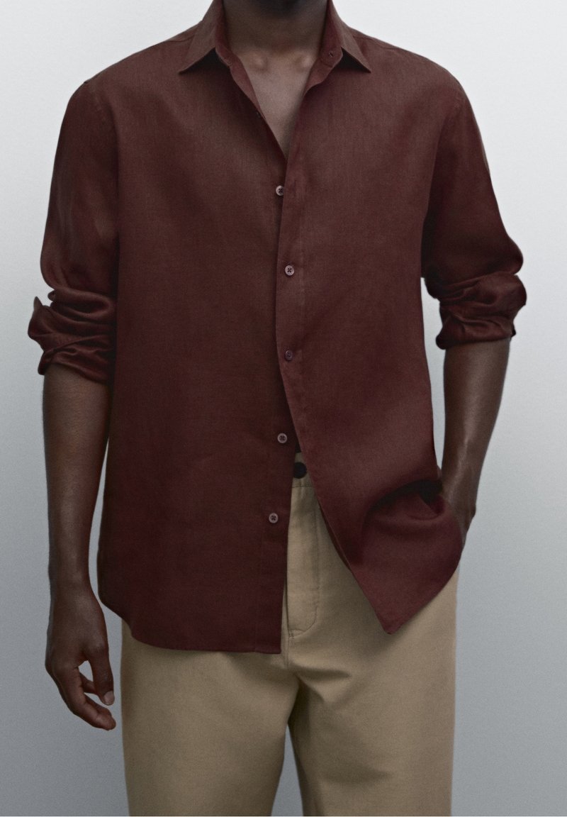Homme portant une chemise marron foncé à manches longues déboutonnée avec les manches retroussées et un pantalon beige, debout avec une main dans la poche.