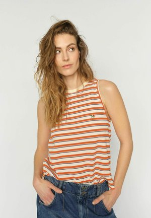 Jeune femme aux longs cheveux ondulés, portant un haut sans manches à rayures orange, beige et noire, un jean bleu, les mains dans les poches, regardant sur le côté.