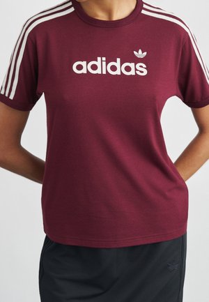 Person wearing a maroon Adidas t-shirt with white stripes on sleeves and Adidas logo text, paired with black Adidas pants.