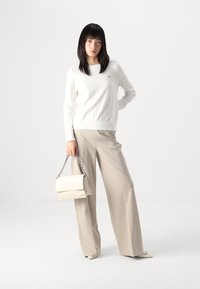 Maglione bianco a maglia, pantaloni beige a gamba larga e una borsa beige chiaro con tracolla a catena. Texture morbide e design minimalista.