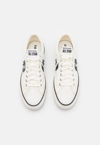 Pár bílých nízkých tenisek Converse All Star s černými hvězdami a bílými tkaničkami, pohled shora na bílém pozadí.