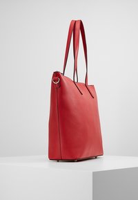 Červená kožená taška na rameno s dvojitými popruhy, stříbrným kovovým kroužkem a strukturovaným obdélníkovým dnem na bílém podstavci.