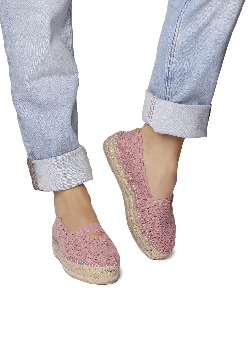 Personne portant un jean bleu clair retroussé et des espadrilles roses au crochet avec des semelles tissées en jute, debout sur fond blanc.