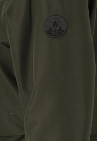 Olivgrön jacka med en texturerad yta, prydd med en svart cirkulär logotyp med bergdesign på den övre ärmen.