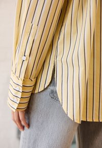 Camicia gialla a righe con linee verticali scure, polsini con bottoni e spacco laterale. Indossata sopra jeans in denim blu. Tessuto liscio e leggero.