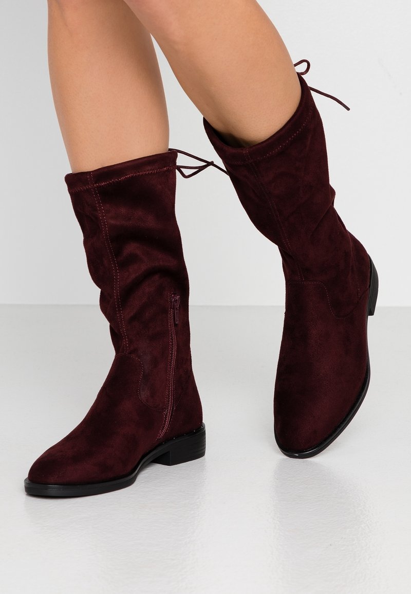Bottes hauteur genou en daim doux couleur bordeaux, avec une fermeture éclair latérale, une semelle plate noire et des liens réglables en haut pour un ajustement parfait.