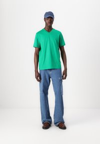 T-shirt en coton vert à col en V et à manches courtes, associé à un jean en denim baggy bleu clair et des chaussures marron. Une casquette bleue complète la tenue.