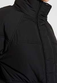 Černá péřová bunda s vysokým límcem, prošívanou strukturou a hladkým vnějším povrchem. Obsahuje viditelné švy a volný střih.