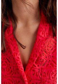 Vêtement en dentelle rouge avec des motifs floraux complexes et un décolleté en V profond, présentant un tissu texturé et une finition douce.