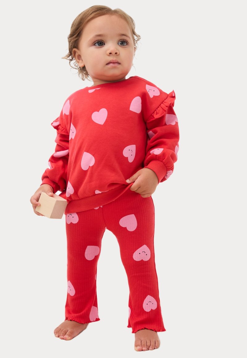 Rød sweatshirt med lyserøde hjerte mønstre og flæseærmer, parret med ribbede røde bukser med matchende hjerte design. Træklods holdes.