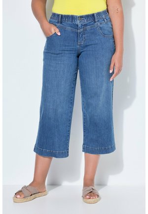 Persona che indossa jeans corti in denim blu con vita elastica, top giallo e sandali beige senza cinturino, su sfondo bianco semplice.