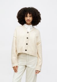 Béžový cropped cardigan vyroben�ý z měkkého pleteného materiálu, s V-výstřihem, žebrovanými manžetami a pěti černými knoflíky na přední straně.